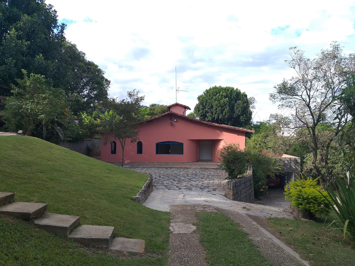 A spacious, single-story house features a warm, terracotta exterior. The building is surrounded by a well-kept lawn and trees, with a stone pathway leading to the entrance. Large windows allow natural light into the interior, providing an inviting appearance.