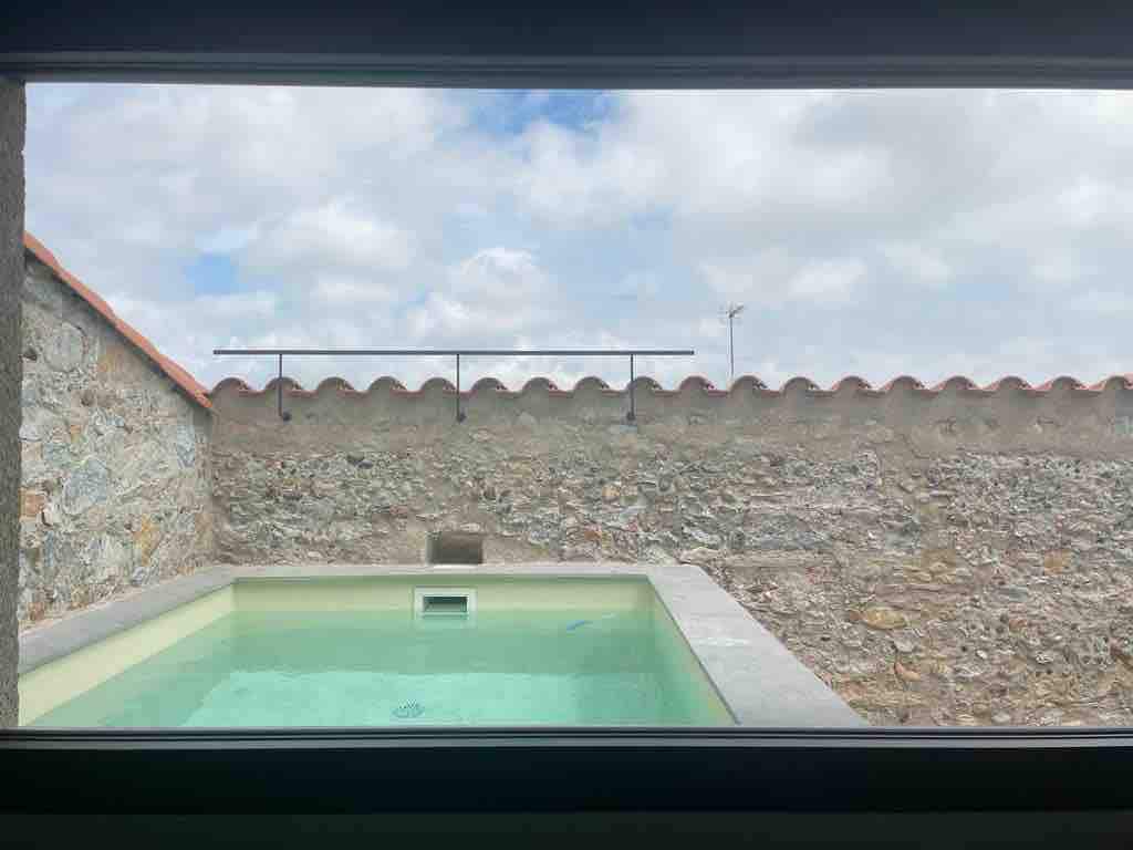 A serene view through a window showcases a refreshing salt-water pool. The pool is surrounded by a smooth concrete border and set against a rustic stone wall topped with traditional clay tiles. The sky above is partly cloudy, creating a calm atmosphere.