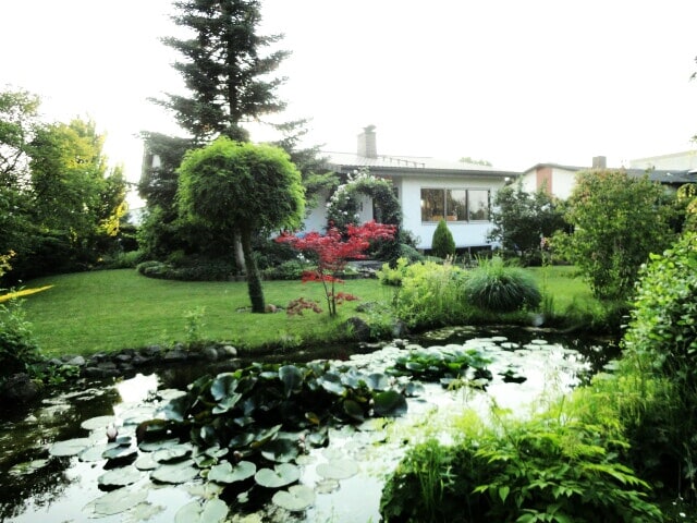 A serene garden scene showcases a lush green lawn framed by various trees and flowering plants. A pond with lily pads is situated in the foreground, reflecting the peacefulness of the surrounding nature, while the bungalow is subtly nestled in the background.