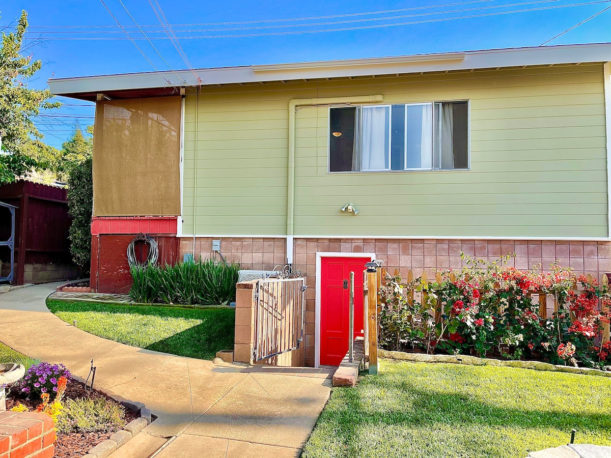 A detached studio welcomes guests with a bright green exterior and a vibrant red door. The pathway, lined with flower beds, leads to the entrance. Large windows illuminate the space, surrounded by well-maintained grass and ornamental plants.
