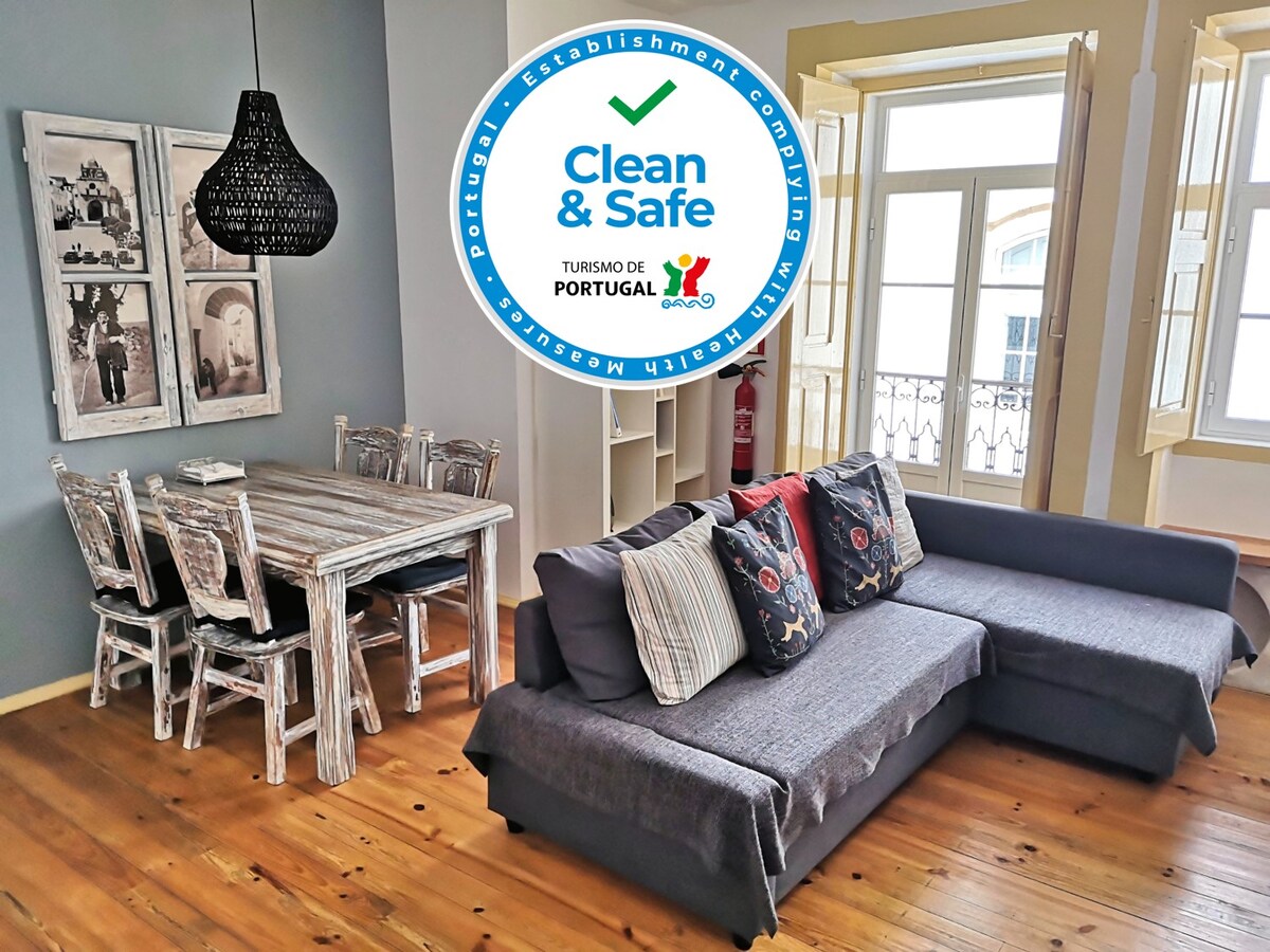 The living area features a comfortable gray sofa with decorative cushions, paired with a wooden dining table and four chairs. Natural light enters through large windows, illuminating the warm wooden floor. A pendant light hangs above the table, enhancing the space's inviting feel.