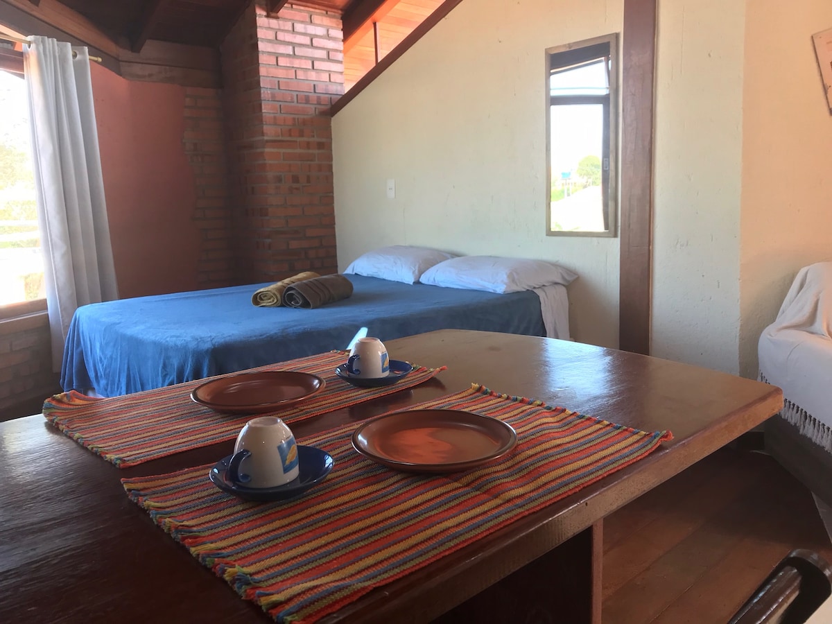 A cozy suite features a spacious bed with a vibrant blue blanket, complemented by soft pillows. A wooden dining table is set with colorful plates and mugs. Natural light filters through a window, illuminating the warm wooden floor and brick walls.