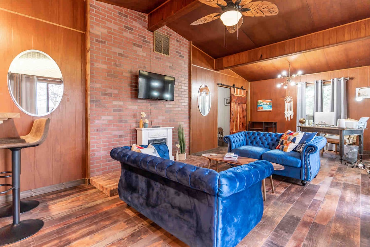 The living room features a cozy blue velvet sofa and a matching armchair, complemented by a wooden coffee table. A wall-mounted television is positioned above an electric fireplace, and large windows allow natural light to fill the space, which is accented by warm wood paneling.