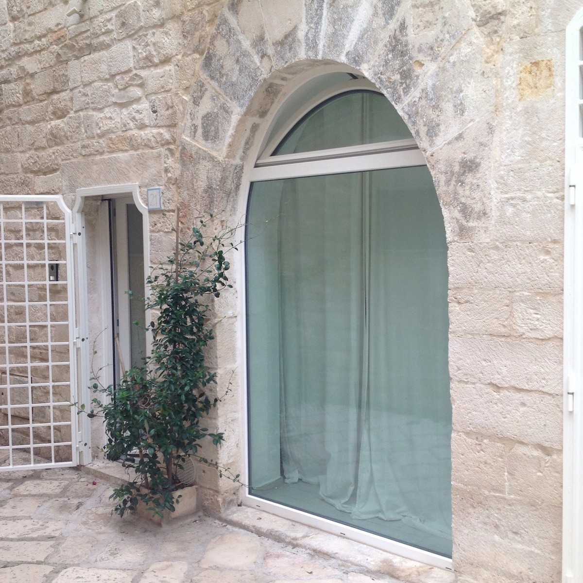 A large arched window showcases sheer curtains, allowing soft light to filter into the space. A small adjoining entrance is visible, framed by a simple white gate. A modest green plant adds a touch of nature to the stone wall backdrop.