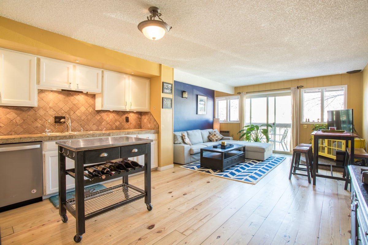The open living area showcases hardwood flooring and a combined kitchen and sitting space. A cozy sofa is placed against a blue wall, with large windows allowing natural light. A kitchen island with wine storage adds functionality, while a small dining table complements the space.