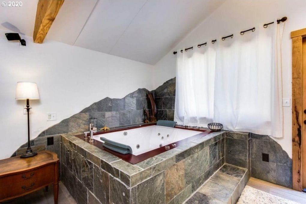 A spacious jetted Jacuzzi is set in a clean, stone-tiled nook, with smooth stone accents creating a natural aesthetic. Soft white curtains frame the window, allowing light to filter in. A side table with a lamp adds subtle illumination to the tranquil space.