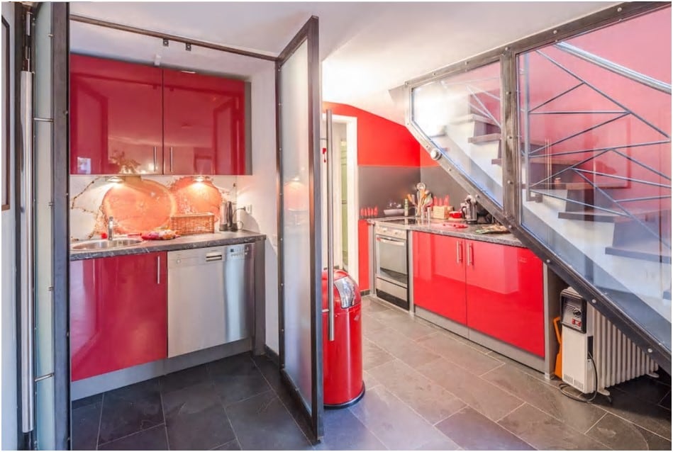 The kitchen features modern red cabinetry with stainless steel appliances, including a refrigerator and dishwasher. A granite countertop provides ample workspace. The floor is covered in sleek black tiles, enhancing the contemporary design. A staircase is visible, leading to the upper level.