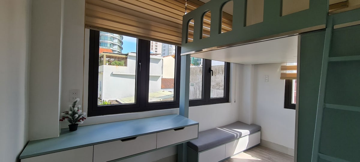 A well-lit room features a cozy lofted sleeping area. Two large windows allow natural light to fill the space. A grey bench runs along the wall, complemented by a minimalist desk with storage drawers below. A small plant adds a touch of greenery.