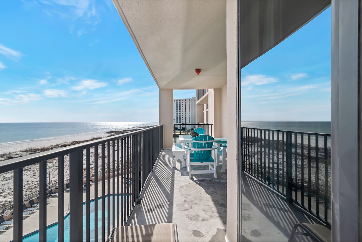 A spacious balcony offers stunning views of the Gulf of Mexico, featuring turquoise chairs and a small table. Bright sunlight reflects off the water, while the beach stretches beyond. The balcony railing creates a contrast against the clear blue sky, inviting relaxation.