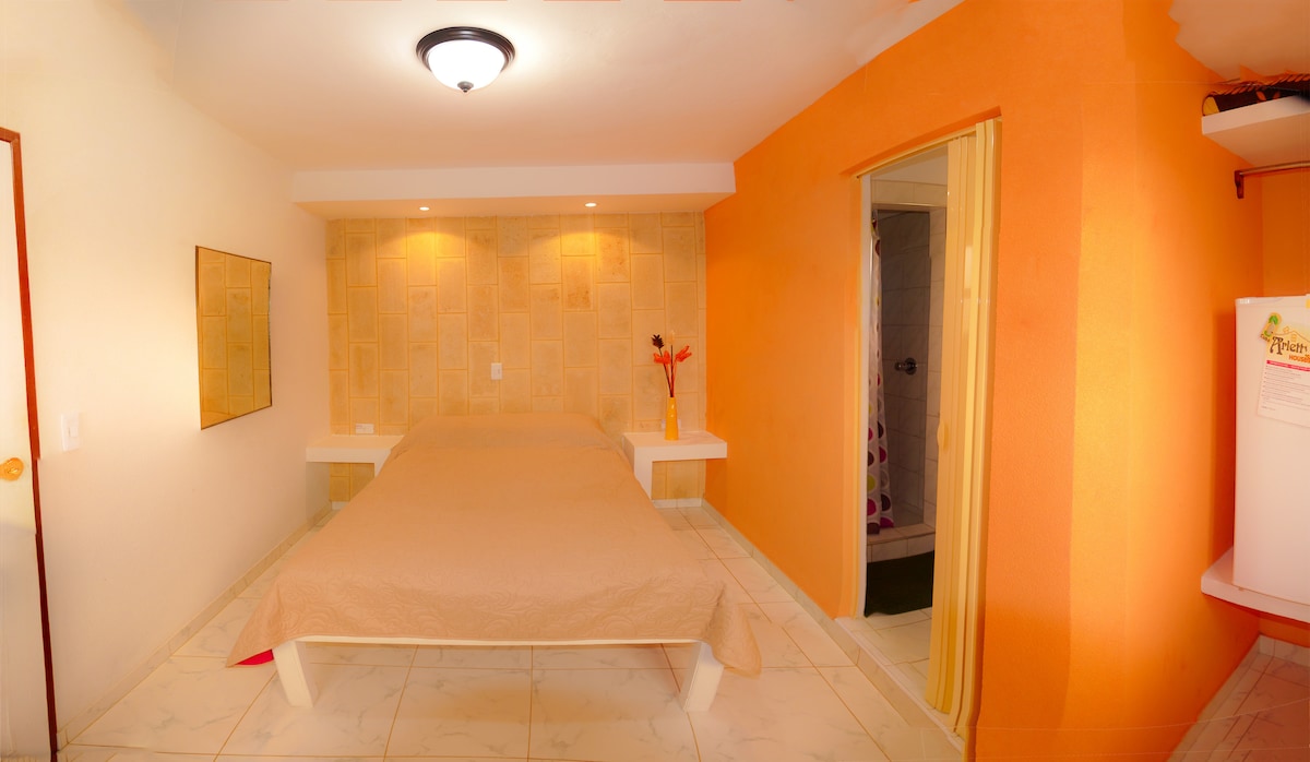 A modern room features a spacious bed with a neutral-colored coverlet. An accent wall in warm orange adds color, while soft lighting enhances the ambiance. A small refrigerator is visible, and a bathroom doorway is seen on the right, providing quick access.