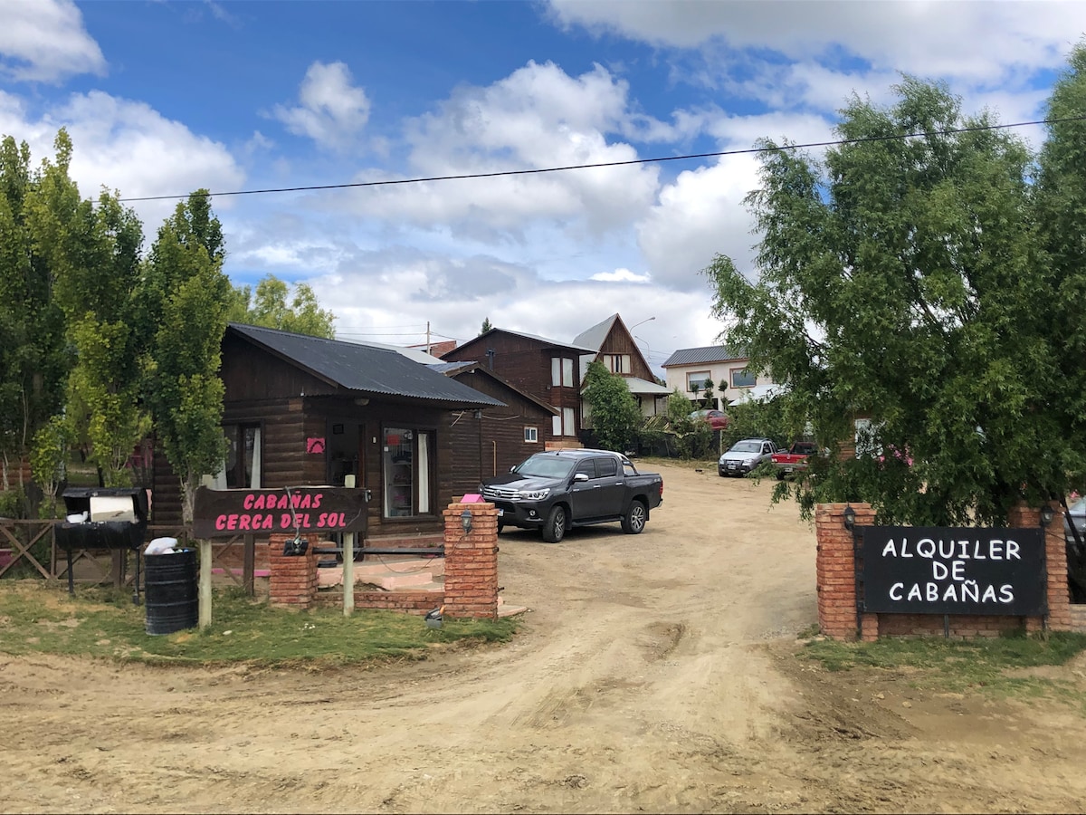 The entrance to the cabin area is shown, featuring wooden structures and a gravel path. Vehicles are parked in designated areas, surrounded by green trees and a clear blue sky. Signage announces cabin rentals, contributing to a welcoming atmosphere.