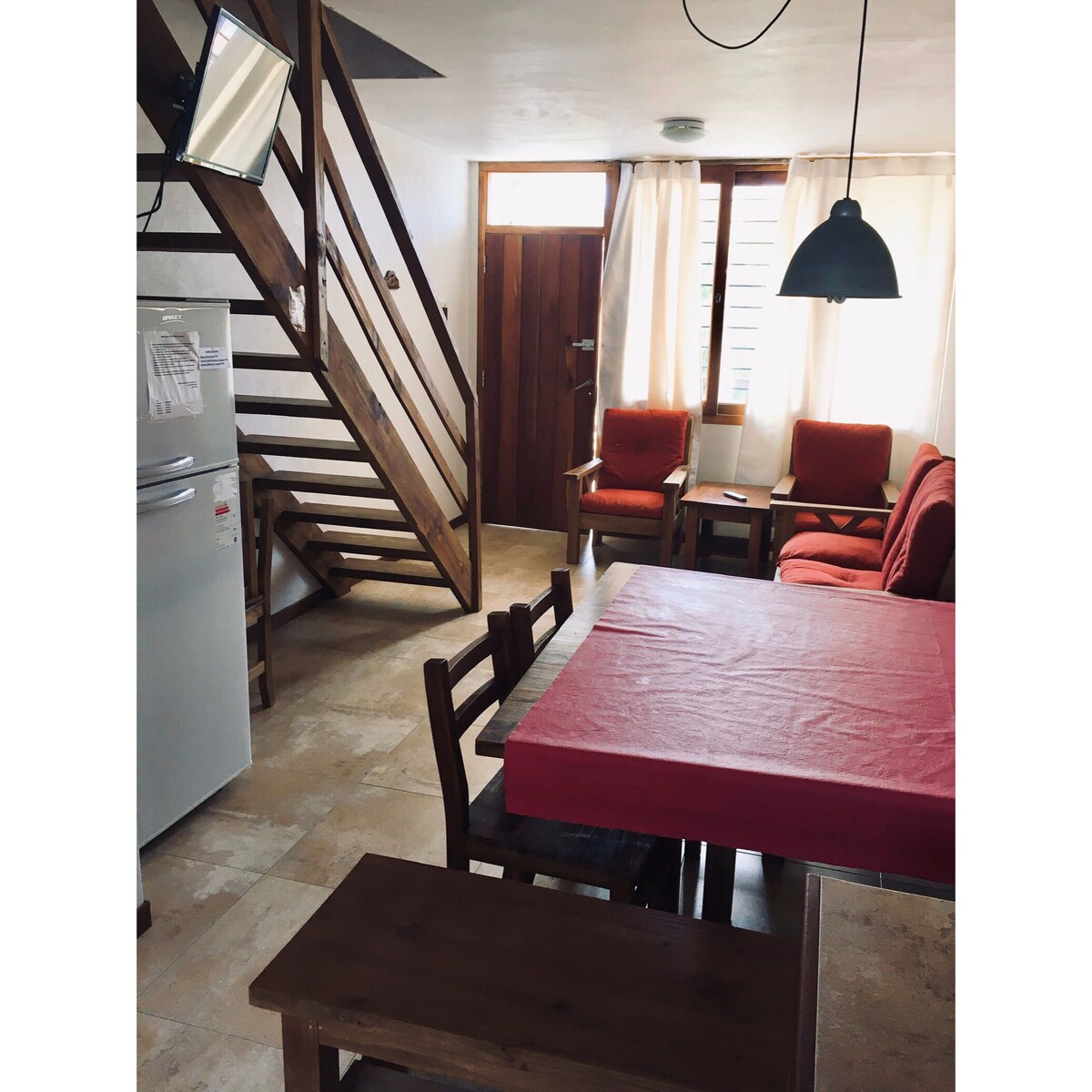 A dining area is visible, featuring a rectangular table with red tablecloth, surrounded by wooden chairs. In the background, a comfortable sitting area includes two red chairs and a small coffee table. A staircase leads to the upper level, while natural light enters through sheer curtains.