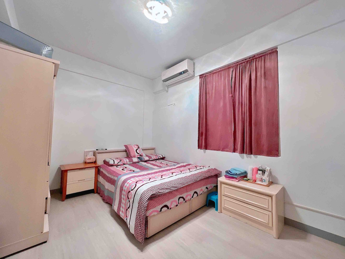 A clean and tidy bedroom is equipped with a spacious bed, air conditioning unit, and light wood furniture. A chest of drawers and bedside table offer ample storage. A window is covered with burgundy curtains, allowing for natural light while maintaining privacy.