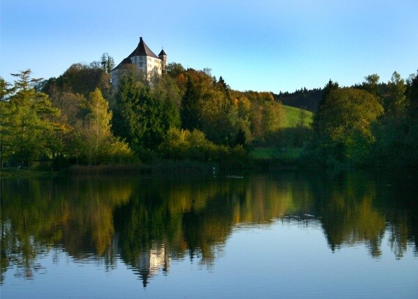 A serene lake mirrors the lush landscape and a historic building perched atop a hill. The scene captures trees in varying shades, reflecting the tranquility of nature during a clear day.