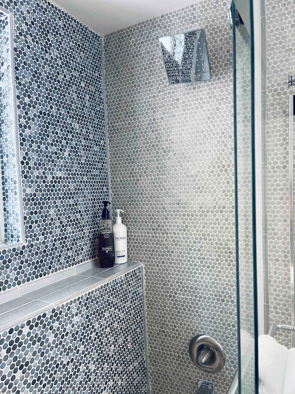 A modern bathroom is depicted, featuring circular mosaic tiles in shades of gray and white. A clear glass shower enclosure is present, along with a built-in shelf displaying toiletries. Natural light enters through a nearby mirrored window, enhancing the contemporary design.