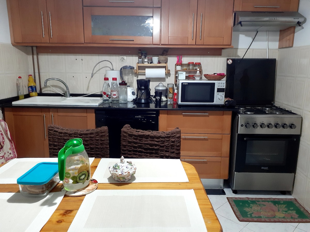 The kitchen features wooden cabinetry with a black countertop. Essential appliances, including a microwave, coffee machine, and stove, are visible. A table is set with a green pitcher and a bowl, providing functional dining space. Nearby items include a dishwasher and an assortment of kitchenware.