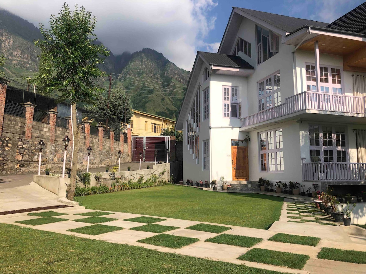 A spacious villa is surrounded by a well-maintained garden, featuring manicured grass and decorative pathways. The exterior showcases large windows and a welcoming entrance. Lush green mountains rise in the background, completing the peaceful and serene environment of the property.