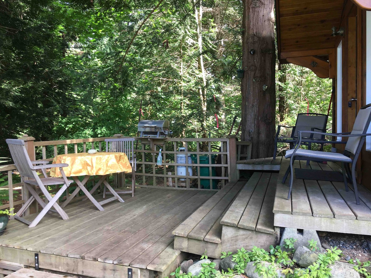 A wooden deck features a rectangular table set for two and a smaller folding table. Comfortable chairs are positioned around the space, with a grill nearby. Lush greenery frames the area, providing a sense of privacy and tranquility.