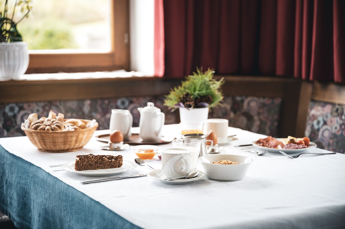 A dining table is set with a white tablecloth, featuring a variety of breakfast items. A basket holds baked goods, accompanied by plates of eggs and cured meats. Coffee and tea are served in porcelain pots and cups, with fresh fruit and a bowl of cereal also visible.
