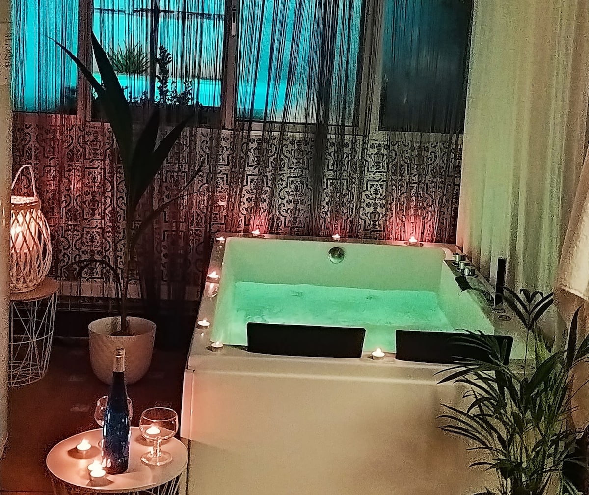 A private jacuzzi is displayed, surrounded by candles and soft ambient lighting. Tropical plants are positioned nearby, enhancing a serene atmosphere. The area features decorative tiles and sheer curtains, creating an intimate space ideal for relaxation.