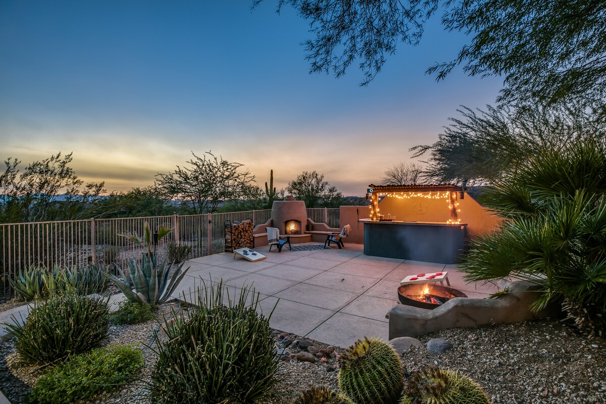 An outdoor space features a covered fireplace, surrounded by desert landscaping and seating areas. Cacti and textured plants enhance the natural scenery, while string lights add a soft glow to the ambiance during twilight. The spacious patio invites relaxation and enjoyment of the serene surroundings.