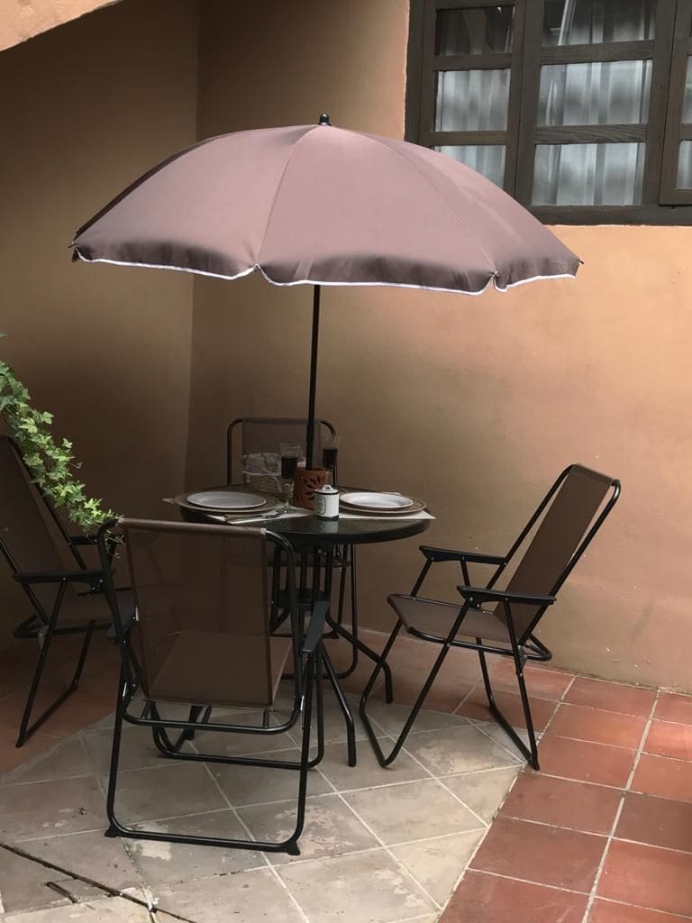 An outdoor dining area is presented with a round black table and four matching chairs. A brown umbrella provides shade over the table, which is set with plates and utensils. The surrounding wall and floor tiles reflect a warm, earthy color.
