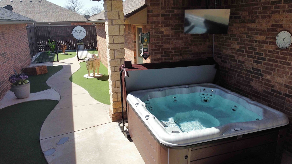 A private Nordic Spa is featured, equipped with jets and a surrounding patio area. A flat-screen TV is mounted nearby, providing entertainment. The space is framed by a walkway, leading to a serene green area with minimal landscaping, enhancing relaxation options.