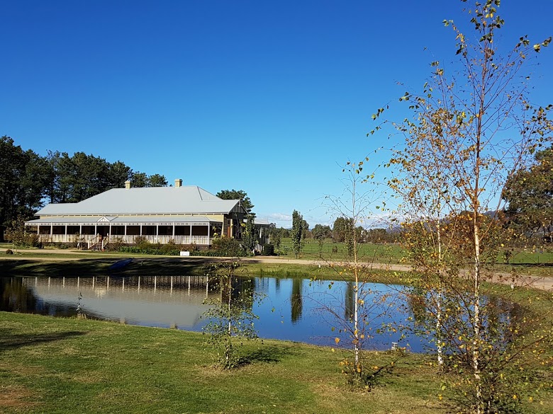 A spacious house is seen beside a tranquil pond, surrounded by lush green grass and trees. The clear blue sky reflects in the water, enhancing the serene countryside landscape. The property’s expansive outdoor area provides a peaceful setting for relaxation.