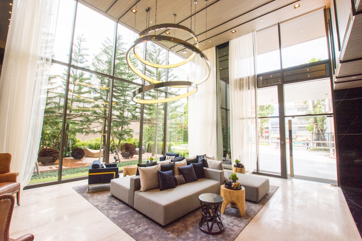 A spacious lobby area is presented with large windows allowing natural light to fill the space. Comfortable seating is arranged in a cozy configuration, complemented by decorative cushions. Modern light fixtures hang from the ceiling, enhancing the inviting ambiance of the room.