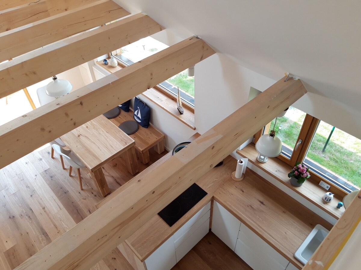 A modern kitchen is viewed from above, featuring wooden beams and light-colored cabinetry. A wooden table with chairs is visible, along with a cooktop and a window bringing in natural light. A plant decorates the countertop, enhancing the welcoming environment.