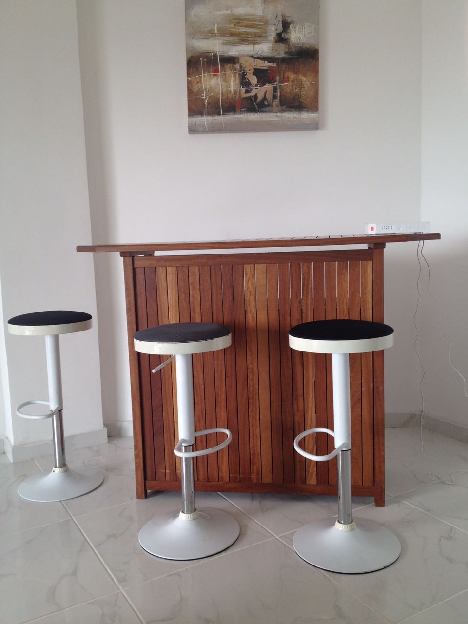 A wooden bar counter is featured against a light wall, accompanied by two modern swivel stools. The stools have white bases and black seating, providing a minimalist design. Above the bar, an abstract artwork adds a touch of color to the space.