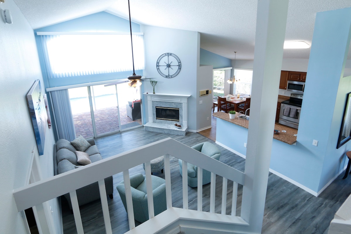 A view from the upper level showcases a spacious living area with a light blue color scheme. A cozy sectional sofa is positioned near a fireplace, while large windows allow natural light to illuminate the space. The dining area and kitchen are visible in the background.