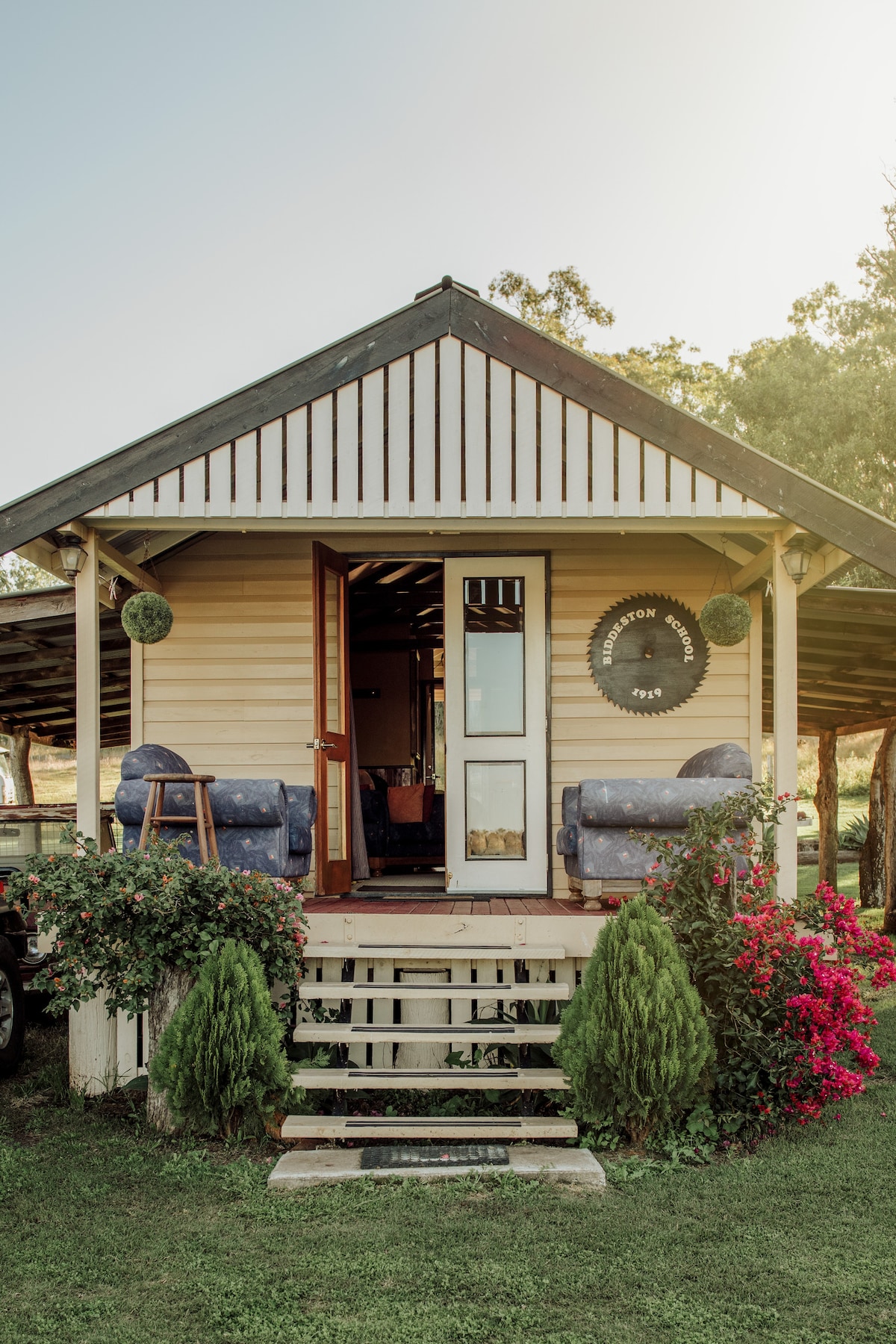 The entrance of the Original Biddeston School is framed by lush greenery and colorful flowers. Two outdoor chairs are positioned on the porch, inviting relaxation. The building features a light-colored exterior with wooden accents and a large door leading to the interior.
