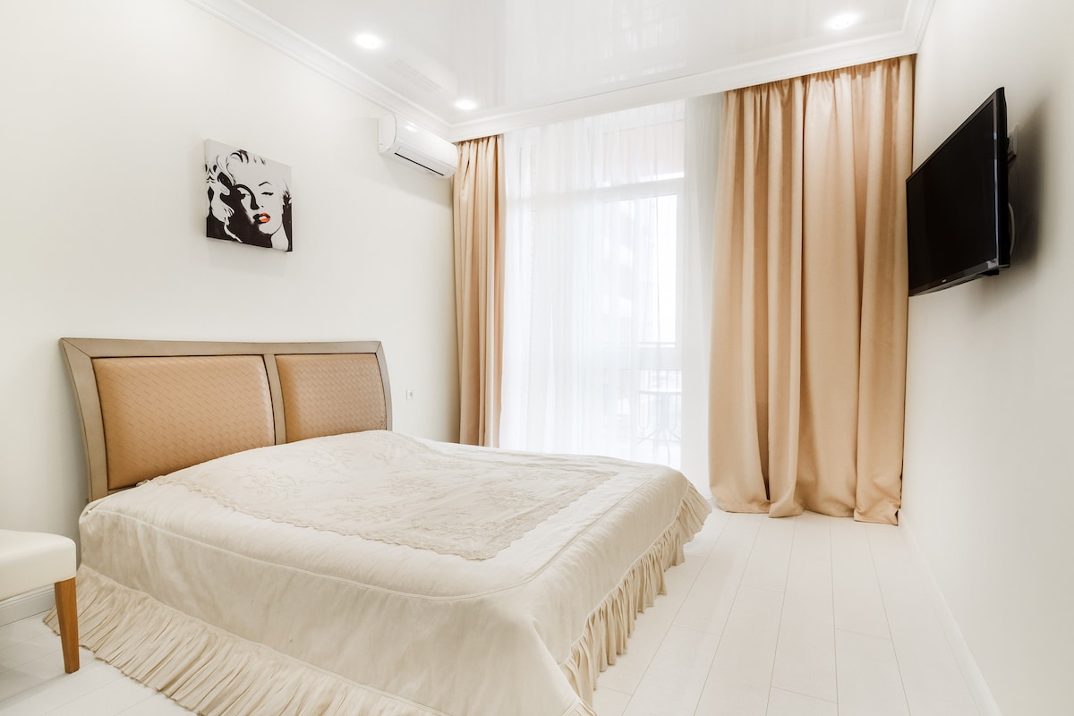 A spacious bedroom features a large bed dressed in a neutral-toned coverlet, with elegant ruffled edges. Natural light enters through floor-to-ceiling curtains, creating a soft ambiance. A mounted television is visible on the opposite wall, alongside minimalistic artwork adding a touch of character.