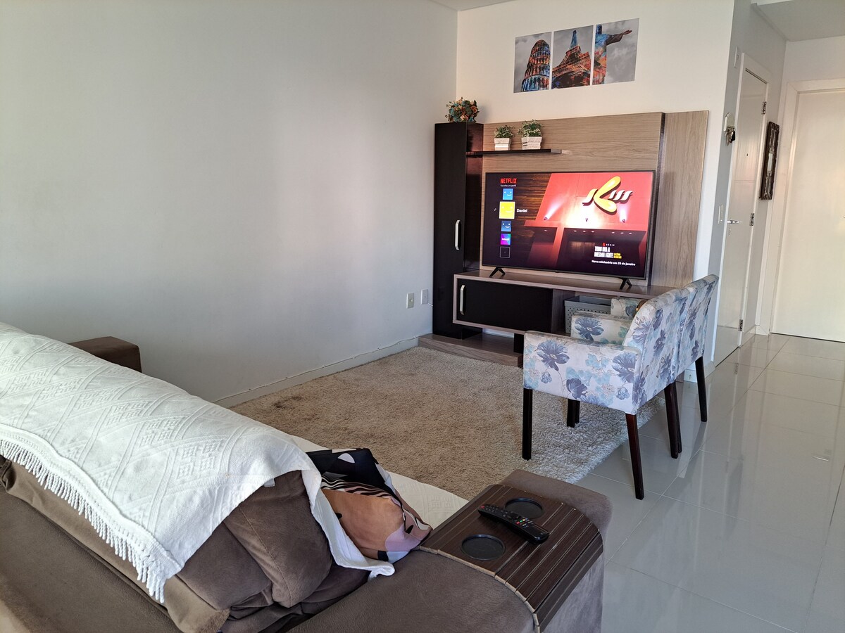 A living area is shown, featuring a brown sofa with light-colored throw blankets and a small patterned chair. A wooden entertainment unit houses a flat-screen TV. A light rug covers the tiled floor, and wall decorations are visible in the background.