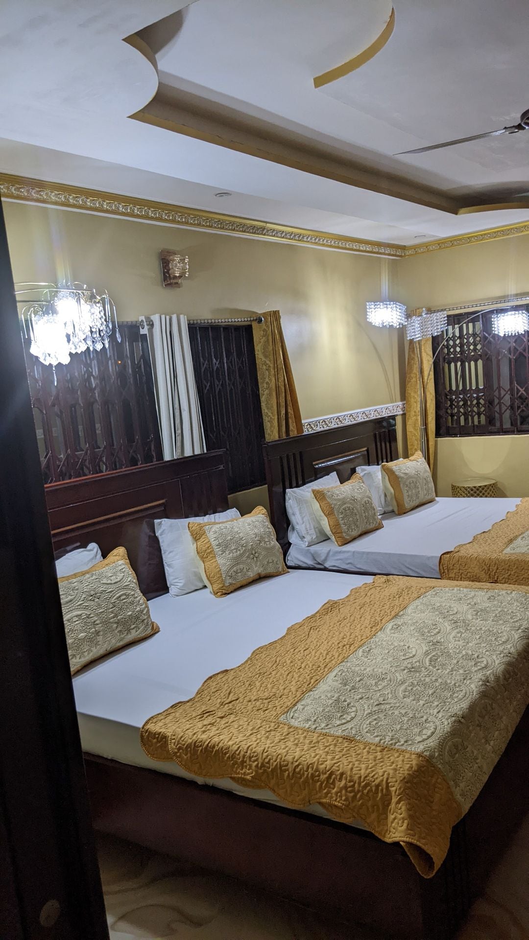 A spacious room features three beds, each adorned with plush pillows and decorative bedding in gold and cream tones. Elegant wall sconces provide soft lighting, while intricate woodwork and patterned curtains enhance the room's character.