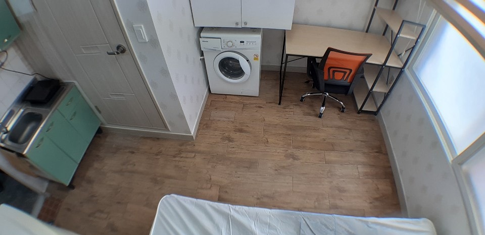 A bright and functional space features a washing machine and a work desk with an orange chair. The kitchen area is equipped with a small stove, and the wooden floor adds warmth to the room. Large windows allow natural light to fill the area.