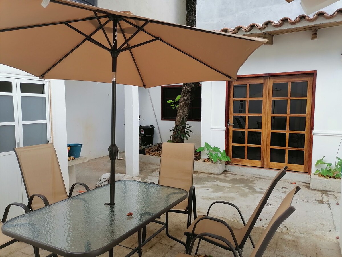 An outdoor seating area is comprised of a glass table surrounded by four chairs beneath a large umbrella. The space features a mix of tiled and concrete flooring, with potted plants and a sturdy wooden door visible in the background.