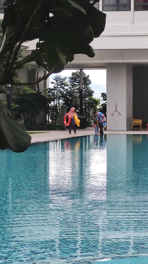 A serene swimming pool is surrounded by lush greenery, reflecting the clear sky. Two individuals are seen approaching the pool's edge, holding colorful flotation devices. The modern architecture creates a relaxing atmosphere, with a shaded area visible nearby.