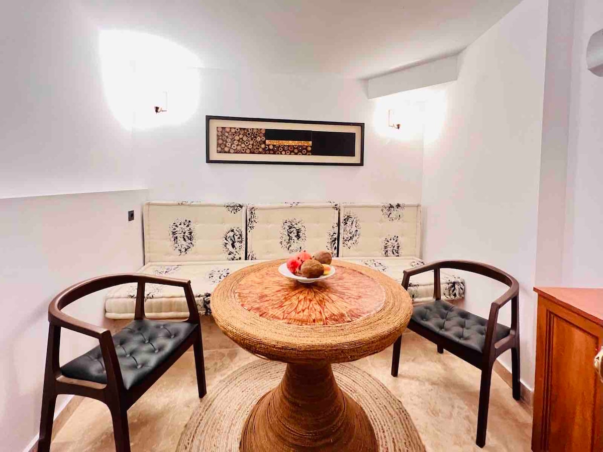 A cozy living area features a circular table at its center, surrounded by two stylish chairs. Light fixtures mounted on the walls provide illumination, while a piece of artwork decorates the wall. A bowl of fruit rests atop the table, adding a touch of color.