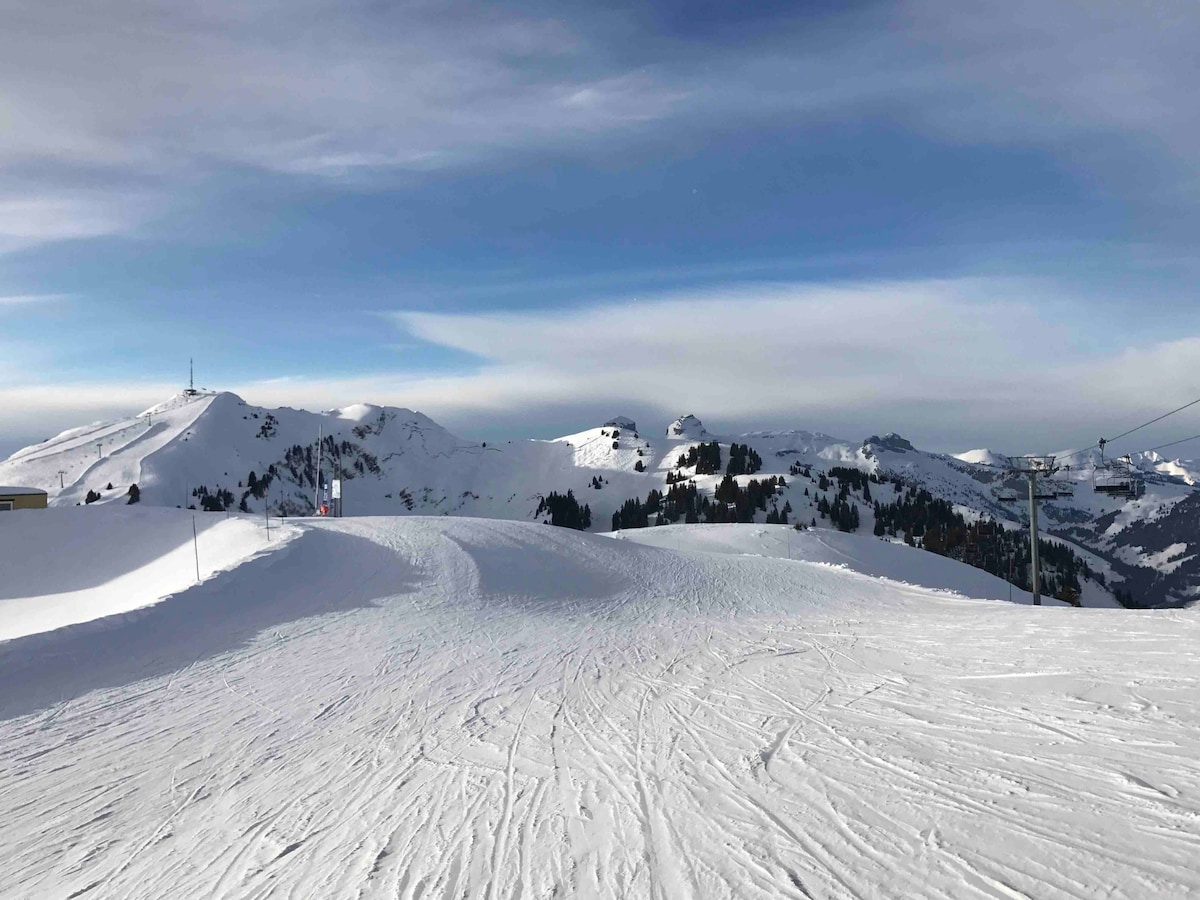 A panoramic view of snow-covered mountains under a clear blue sky is displayed. Smooth, winding ski trails are visible, leading into the serene landscape. Lush green trees peek through the snow, adding contrast to the scene, while distant peaks rise majestically in the background.