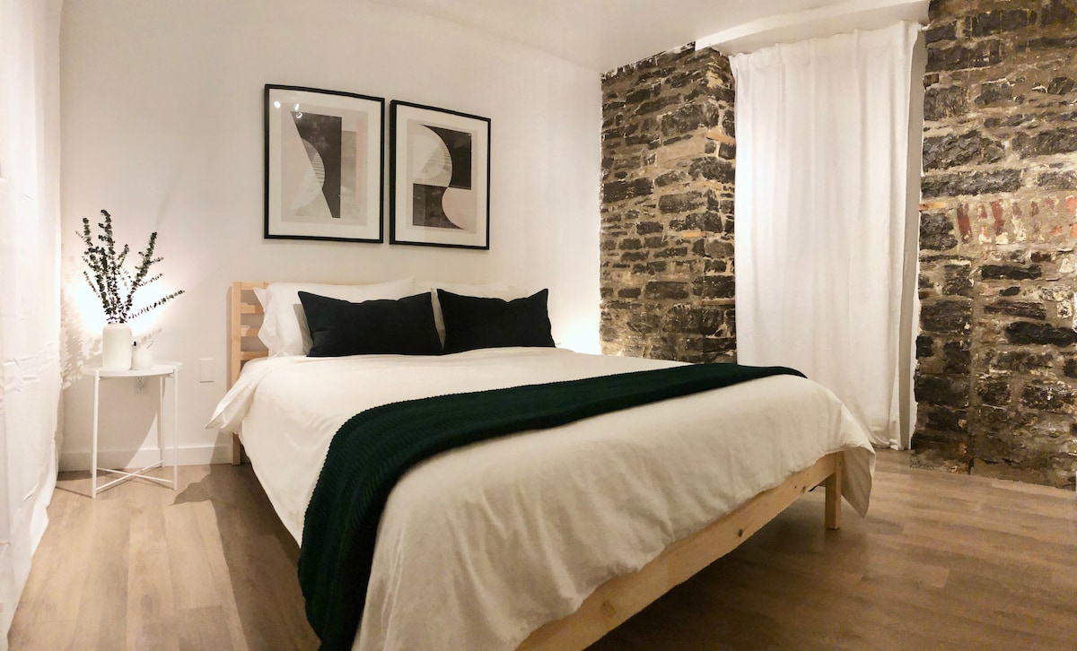 The bedroom features a spacious bed dressed in soft linens, flanked by two framed artworks on the wall. A small white side table holds decorative greenery. Textured stone walls add character, while natural light filters through sheer curtains, enhancing the room's inviting atmosphere.