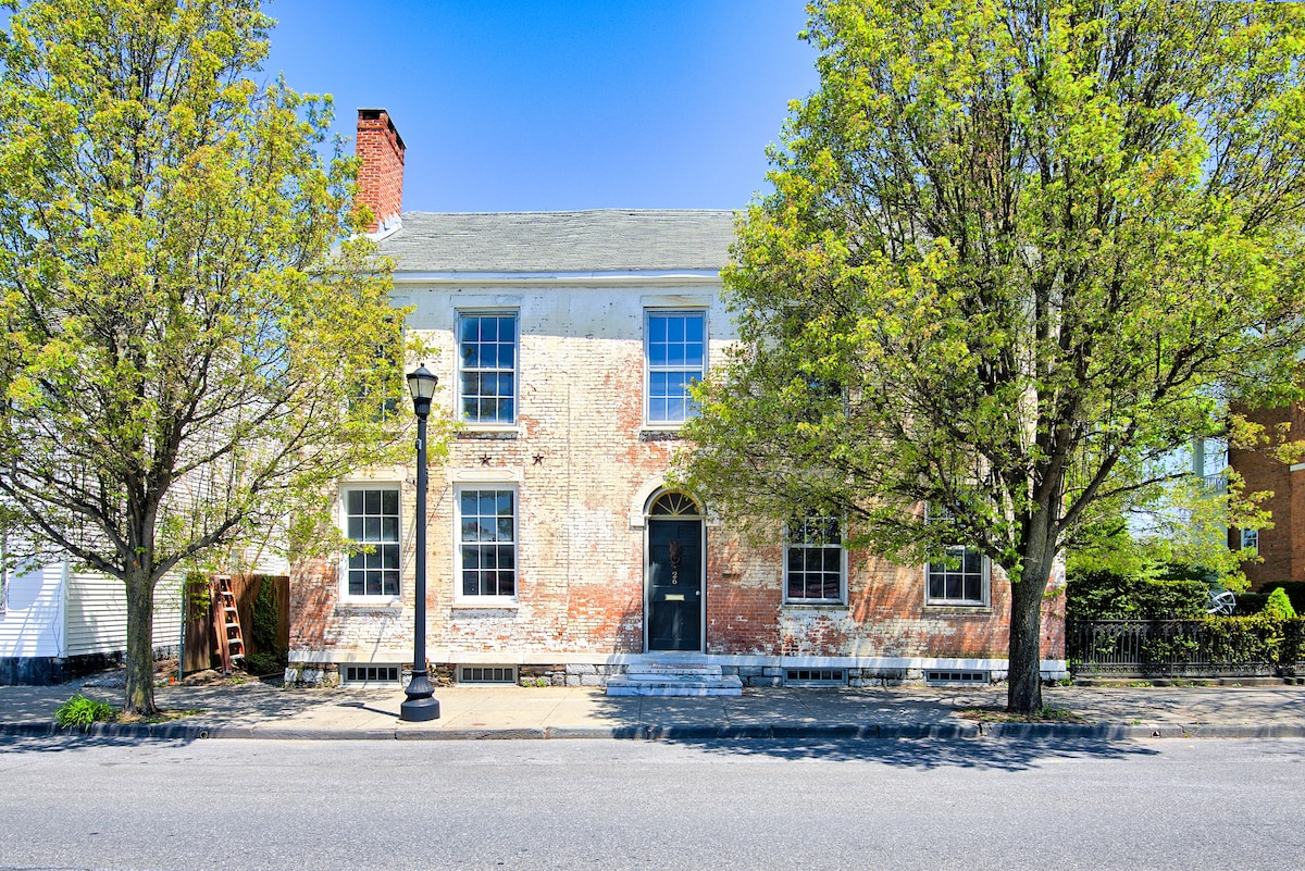 A historic building with a weathered brick facade is surrounded by mature trees. Large windows offer a glimpse inside, while a black door provides entrance. A lamppost stands on the sidewalk, suggesting a serene neighborhood ambiance. The clear blue sky enhances the inviting setting.