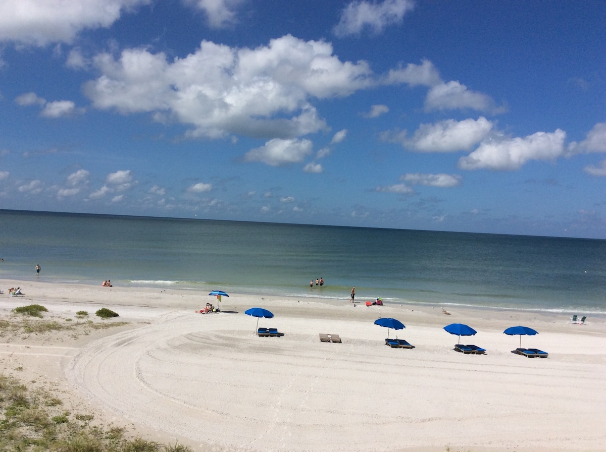 A sandy beach is visible, featuring several blue umbrellas and lounge chairs arranged along the shoreline. Gentle waves can be seen in the calm water, while fluffy white clouds float in the blue sky above, creating a serene coastal atmosphere.