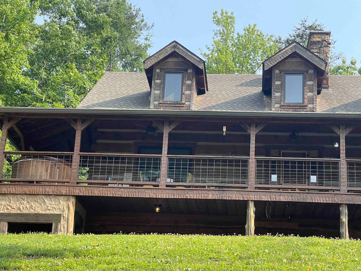 A charming cabin with rustic wooden architecture features a spacious porch supported by stone pillars. Large windows provide light and views, while the surrounding greenery enhances the tranquil setting. A hot tub is visible on the porch, inviting relaxation in nature.