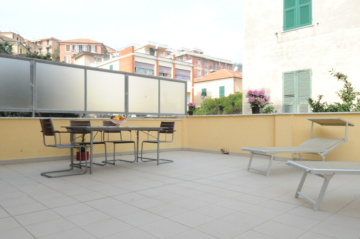 A spacious terrace is presented, featuring a table surrounded by four chairs and two sun loungers. Planters with greenery are positioned along the edges, while semi-transparent panels provide partial enclosure. A view of nearby buildings is visible in the background.