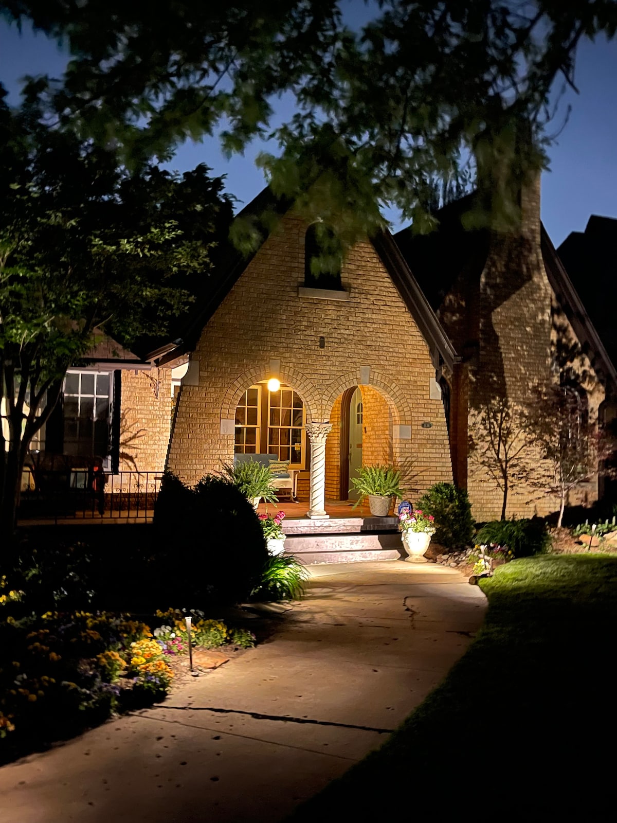 An inviting entrance is highlighted by warm lighting, revealing a charming brick façade adorned with greenery. A path lined with flowers leads to the arched doorway, which is flanked by well-kept plants. The serene ambiance is enhanced by the gently illuminated surroundings.