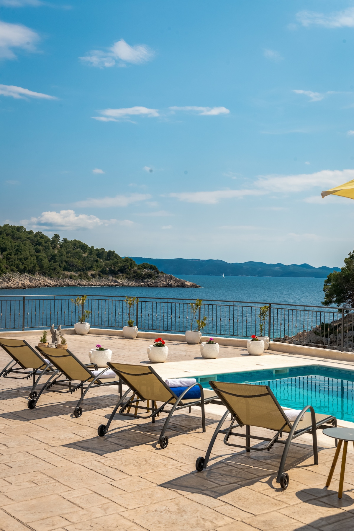 A spacious terrace features multiple sun loungers arranged around a private swimming pool. Potted plants add greenery, and the ocean extends in the background under a clear blue sky, complemented by distant hills and a gentle breeze.