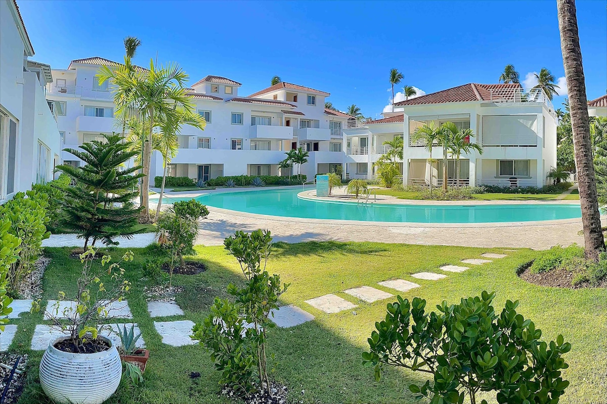 The image captures a serene pool area surrounded by well-maintained greenery. Palm trees and tropical plants are placed around the pool, with a spacious lawn and pathway leading to the inviting turquoise water. The buildings are modern and feature multiple balconies, enhancing the property’s charm.