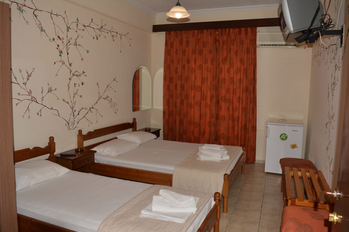 A room with three beds is provided, featuring wooden frames and white linens. Soft orange curtains complement the floral wall design. A small refrigerator, a mirror, and a wooden bench are visible, enhancing the functional space while maintaining simplicity.